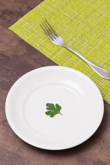 Single one parsley green leaf on white empty plate and fork on table. Diet, fasting, vegan, vegetarian, healthy food concept