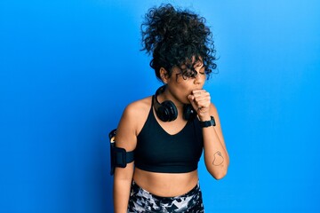 Young hispanic woman wearing gym clothes and using headphones feeling unwell and coughing as...