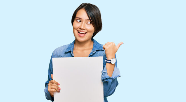 Beautiful young woman with short hair holding blank empty banner pointing thumb up to the side smiling happy with open mouth
