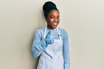 African american woman with braided hair wearing cleaner apron and gloves doing happy thumbs up...
