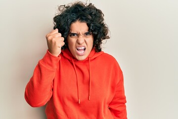 Fototapeta na wymiar Young hispanic woman with curly hair wearing casual sweatshirt angry and mad raising fist frustrated and furious while shouting with anger. rage and aggressive concept.