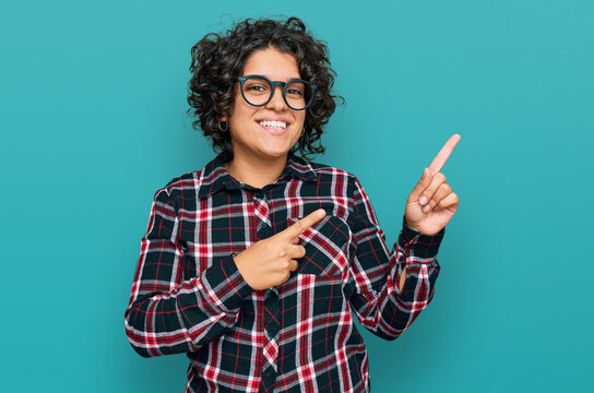 Young hispanic woman with curly hair wearing casual clothes and glasses smiling and looking at the camera pointing with two hands and fingers to the side.