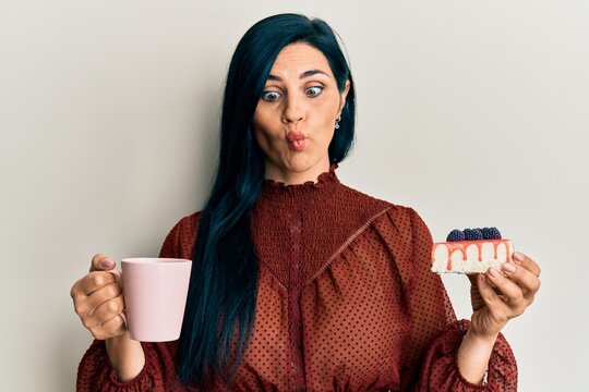 Young caucasian woman holding cup of coffee and cake slice making fish face with mouth and squinting eyes, crazy and comical.
