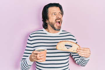 Middle age caucasian man holding bread loaf with heart shape drinking coffee angry and mad screaming frustrated and furious, shouting with anger. rage and aggressive concept.