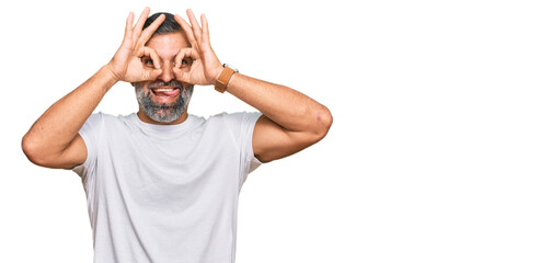 Middle age handsome man wearing casual white tshirt doing ok gesture like binoculars sticking tongue out, eyes looking through fingers. crazy expression.