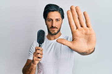 Young hispanic man styling hair using comb with open hand doing stop sign with serious and confident expression, defense gesture
