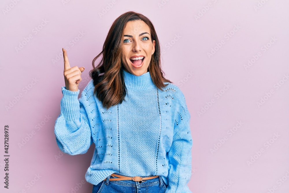 Wall mural Young brunette woman wearing casual winter sweater over pink background pointing finger up with successful idea. exited and happy. number one.