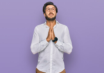 Young hispanic man wearing business shirt and glasses begging and praying with hands together with...