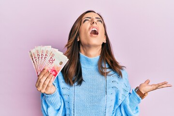 Young brunette woman holding 20 israel shekels banknotes crazy and mad shouting and yelling with aggressive expression and arms raised. frustration concept.