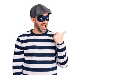 Young handsome man wearing burglar mask smiling with happy face looking and pointing to the side with thumb up.