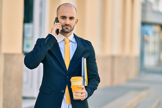 Young hispanic bald businessman with serious expression talking on the smartphone drinking coffee at the city