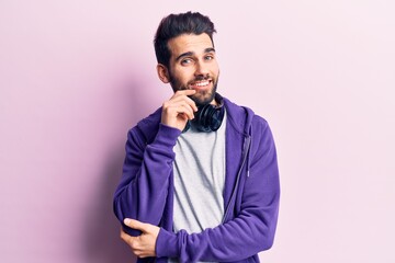 Young handsome man with beard listening to music using headphones smiling looking confident at the camera with crossed arms and hand on chin. thinking positive.