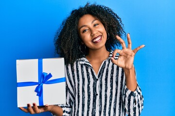 Beautiful african american woman with afro hair holding gift doing ok sign with fingers, smiling...