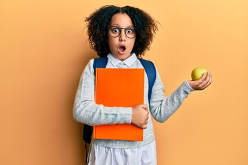 Young little girl with afro hair wearing school bag holding books and green apple afraid and shocked with surprise and amazed expression, fear and excited face.