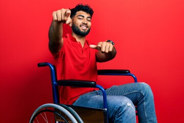 Arab man with beard sitting on wheelchair pointing to you and the camera with fingers, smiling...
