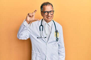 Middle age indian man wearing doctor coat and stethoscope smiling and confident gesturing with hand doing small size sign with fingers looking and the camera. measure concept.