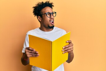 Young african american man with beard reading a book wearing glasses angry and mad screaming frustrated and furious, shouting with anger. rage and aggressive concept.