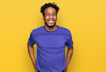 Young african american man with beard wearing casual purple t shirt winking looking at the camera...