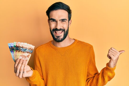Young hispanic man holding canadian dollars pointing thumb up to the side smiling happy with open mouth