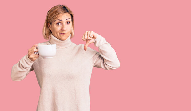 Young blonde woman holding coffee with angry face, negative sign showing dislike with thumbs down, rejection concept