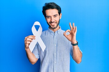 Young hispanic man holding blue ribbon doing ok sign with fingers, smiling friendly gesturing...