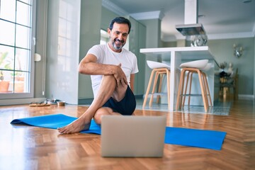 Middle age man with beard training and stretching doing exercise at home looking at sport video on...