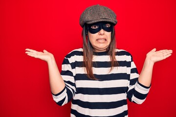 Young beautiful brunette burglar woman wearing cap and mask over isolated red background clueless and confused with open arms, no idea and doubtful face.