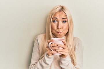 Young blonde woman drinking a cup of coffee puffing cheeks with funny face. mouth inflated with air, catching air.