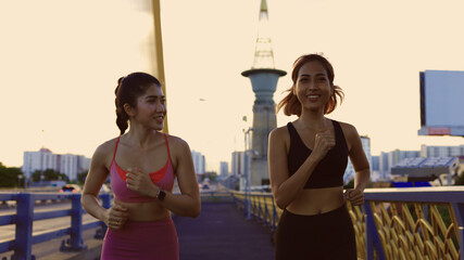 Two healthy and fit young female friends jogging on city street during early morning while talking and smiling in city