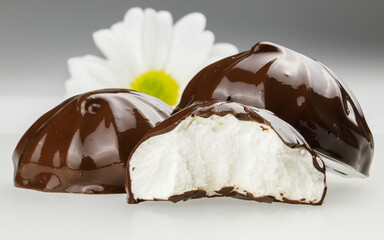 A macro photo of a group of three marshmallows covered in shiny chocolate, one of which is broken in half against the backdrop of white chrysanthemum, which is out of focus.