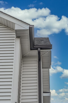 Closeup View Of Dark Metal Gutter System On White Horizontal Vinyl Siding, Fascia, Drip Edge, Soffit, On A Pitched Roof Attic At A Luxury American Single Family Home Cloudy Blue Sky Background