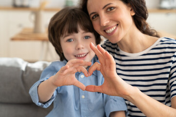 Mom and son making heart hand gesture together happy smiling look in camera at home sitting on sofa. Cute preschool boy and mother take funny family picture photo. Love and happy parenthood concept