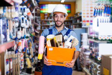 Satisfied adult foreman standing near racks in paint store holding basket with tools
