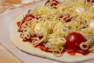 Raw pizza on baker paper on the wooden table. Preparing homemade uncooked cheese pizza with tomato sauce, tomato cherry and dough. 