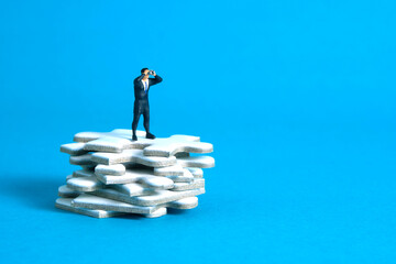 Businessmen standing above jigsaw puzzle piece stack using binoculars above wooden table. Miniature...
