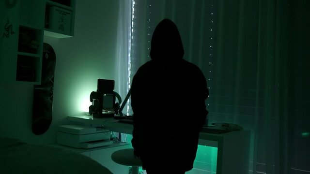 a criminal hackergirl with black hood using laptop and big computers to hack network system from her dark hacker neon room.