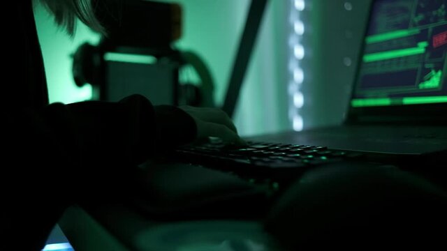 a criminal hackergirl with black hood using laptop and big computers to hack network system from her dark hacker neon room.
