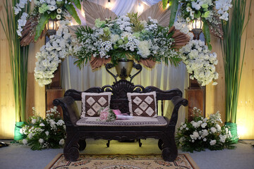 The wedding chair for the bride and groom is made of wood, equipped with two small cushions for the backrest and floral decorations around it.