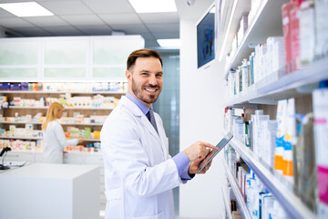 Naklejka na ściany i meble Pharmacist standing by the shelves with medicines and typing on tablet in drug store.
