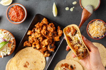 Closeup image of a Mexican taco dish with corn tortillas, precooked, preseasoned chicken pieces, cabbage slaw, shredded cheese, salsa and cream sauce and lime slices. A woman is holding a made wrap.