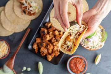 Closeup image of a Mexican taco dish with corn tortillas, precooked, preseasoned chicken pieces, cabbage slaw, shredded cheese, salsa and cream sauce and lime slices. A woman squeezing lemon on wrap.