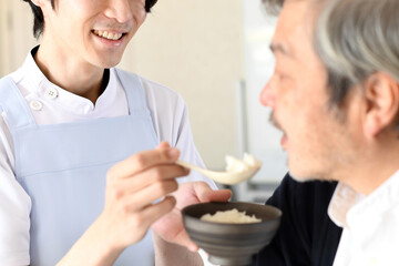 シニア男性に食事の介助をするエプロン姿の若い男性
