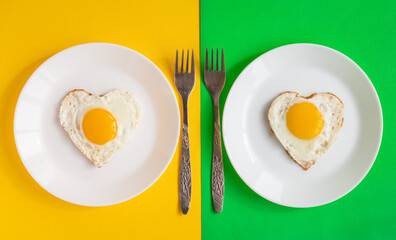 top view of two heart-shaped scrambled eggs, symbol of love, a pair of fried eggs on white plates on yellow and green background, romantic minimalistic template, healthy breakfast for sweetheart