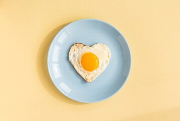 top view of heart-shaped scrambled eggs, symbol of love, single fried egg on blue plate on yellow background, romantic minimalistic template for healthy breakfast
