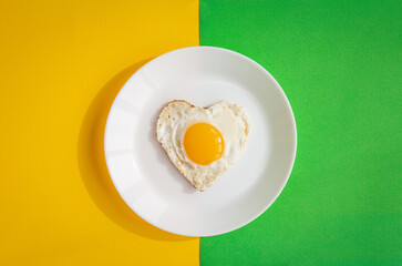top view of heart-shaped scrambled eggs, symbol of love, single fried egg on white plate on yellow and green background, romantic minimalistic template for healthy breakfast