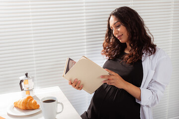 Positive pregnant woman reads book during morning breakfast with coffee and croissants. Concept of pleasant morning and expectation of meeting baby