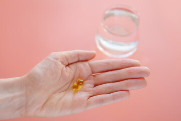 Vitamins of yellow color in a hand on a pink background. Behind - a glass of water. The concept of taking vitamins, dietary supplements, health promotion.