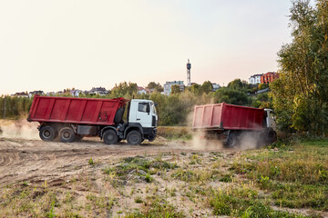 Fototapeta premium Two red dump trucks simultaneously lifted the bodies to unload the sand. Cargo transportation services. Large multi-ton truck. Unloading cargo. Construction site and machinery. Banner. Common view