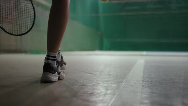 female tennis player is walking on indoor court, closeup of feet shod sporty sneakers, throwing ball, rear view