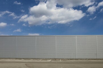 Fototapeta na wymiar Aluminium cladding wall with horizontal stripes under a blue sky with fluffy clouds. Sidewalk, parking and asphalt road in front. Background for copy space